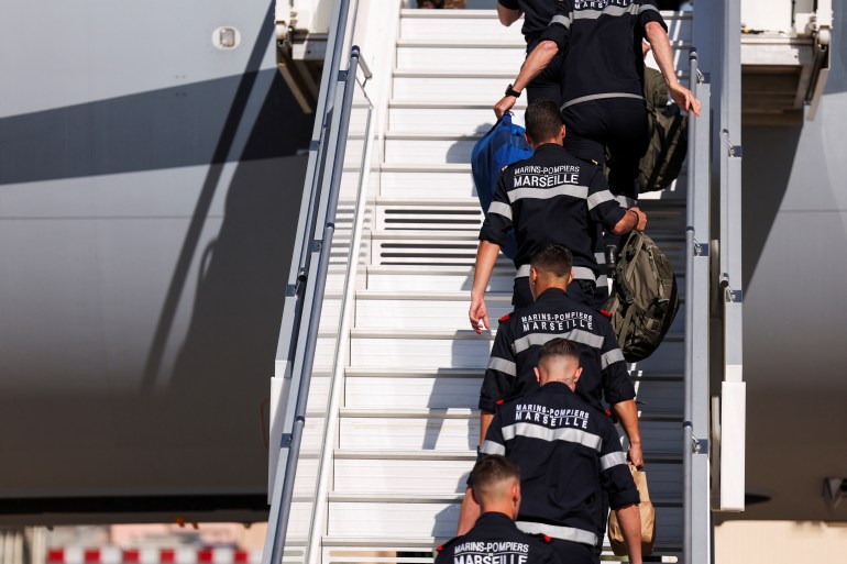 Members of the Marseille Naval Fire Battalion walking up the steps to board a plane. They are wearing blue uniforms with 'Marins Pompiers, Marseilles' written on the back across their shoulders.